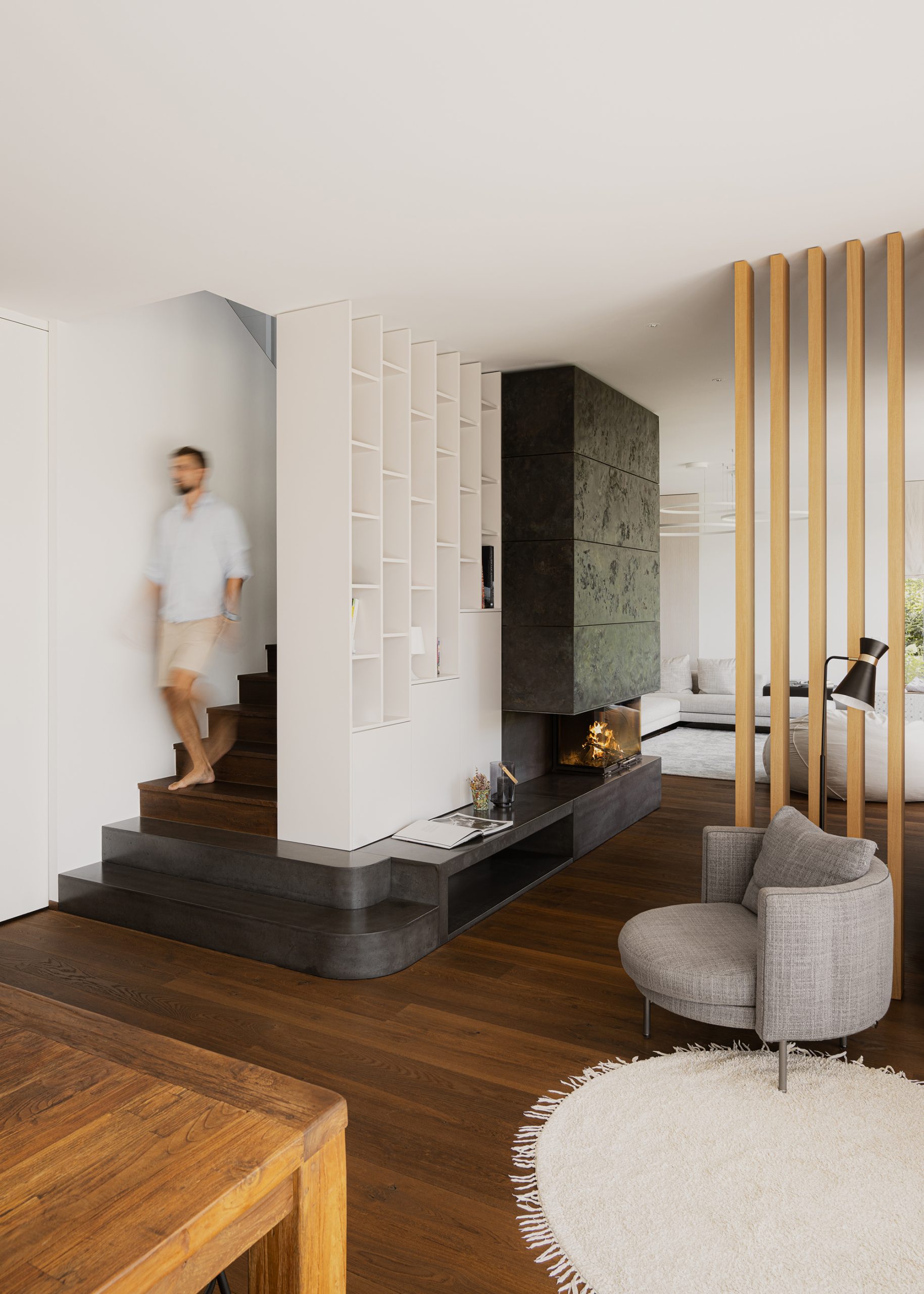 Wohnbereich aus einer Perspektive aus der man die Treppen sehen kann die hinter dem großen, zentralen Kamin nach oben führen und in der unteren linken Ecke ist der Esstisch zu erkennen. Auf der rechten Seite ist ein Raumtrenner aus Holzstäben, welcher Wohn- und Esszimmer optisch voneinander trennt