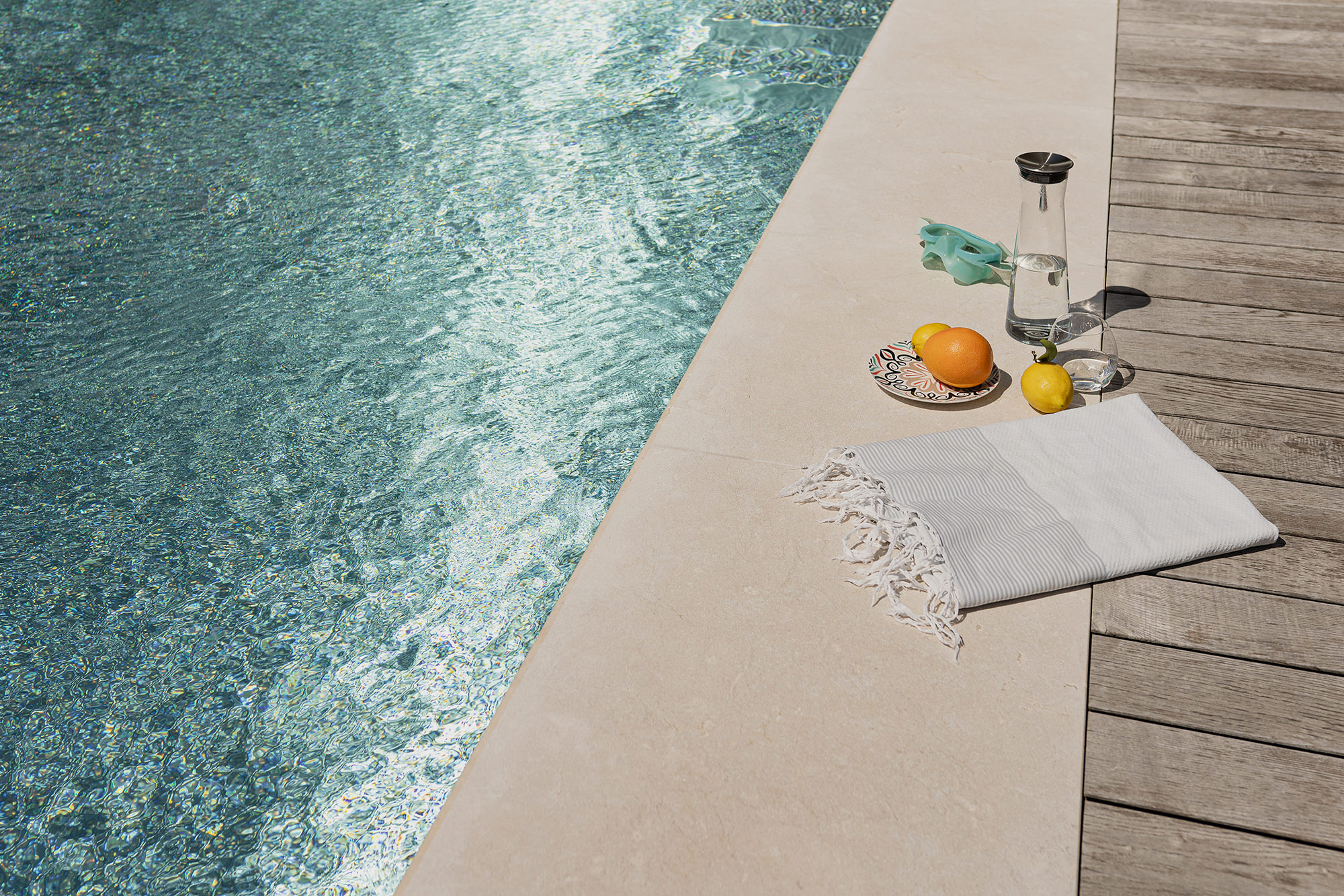 Am Rand eines Outdoor-Pools liegen ein Handtuch, eine Taucherbrille, ein Teller mit Früchten sowie eine Karaffe Wasser mit Glas.