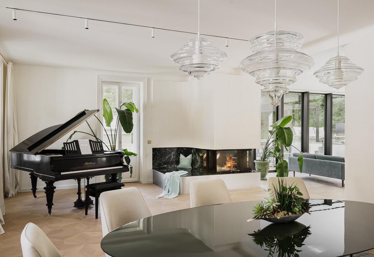 Blick vom Esszimmer auf einen geöffneten schwarzen Flügel links im Raum. In der Mitte steht ein weiß verkleideter Kamin mit einer kleinen Sitzfläche aus Marmor. Rechts ist die Rückseite einer Couch zu sehen.