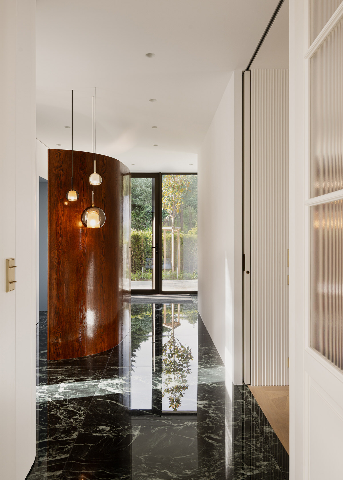 Raum mit dunklen Marmorfliesen, die zur Küche passen, und direktem Blick in den Garten. In der Raummitte steht eine abgerundete Trennwand aus dunklem, rötlichem Holz.