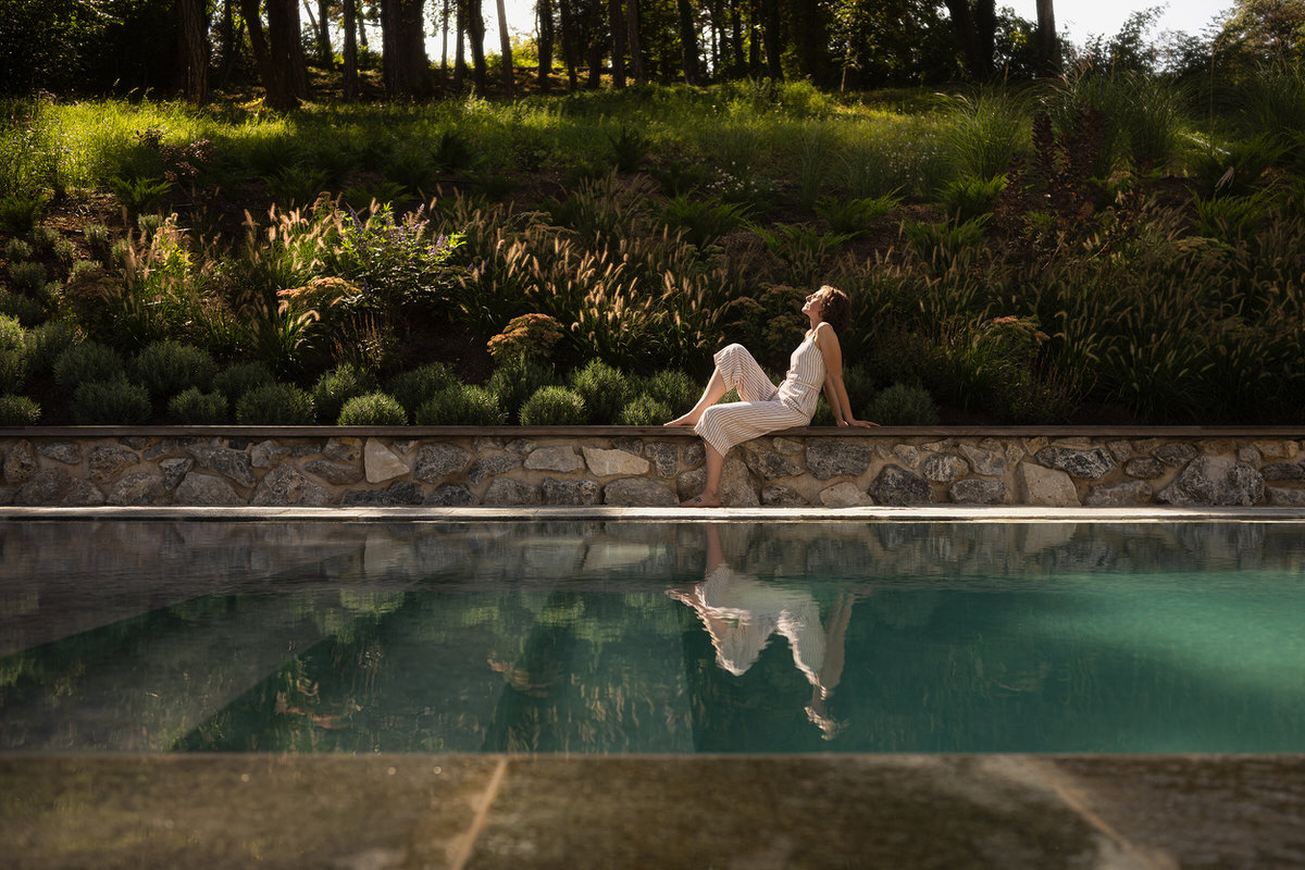 Eine Frau genießt die Sonne neben dem Pool. Im Hintergrund ist ein grün bepflanzter Hang.