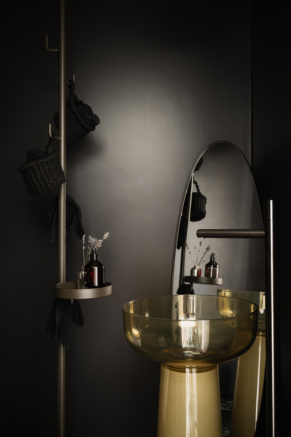 Gläsernes, gelblich getöntes Standwaschbecken mit silberner Bodenarmatur. Dahinter steht ein großer ovaler Spiegel an der Wand. Daneben befindet sich eine deckenhohe Handtuchhalterung mit kleiner Ablage.