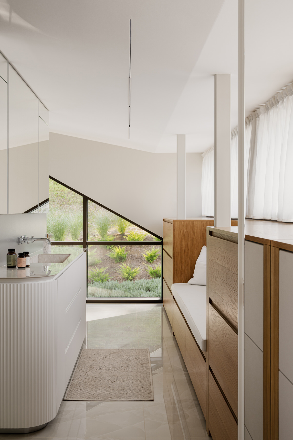 Helles Badezimmer mit einem kleinen Waschbecken auf der linken Seite und einem Holzverbau mit integrierter Sitzgelegenheit gegenüber. Im Hintergrund eine große Fensterfront.