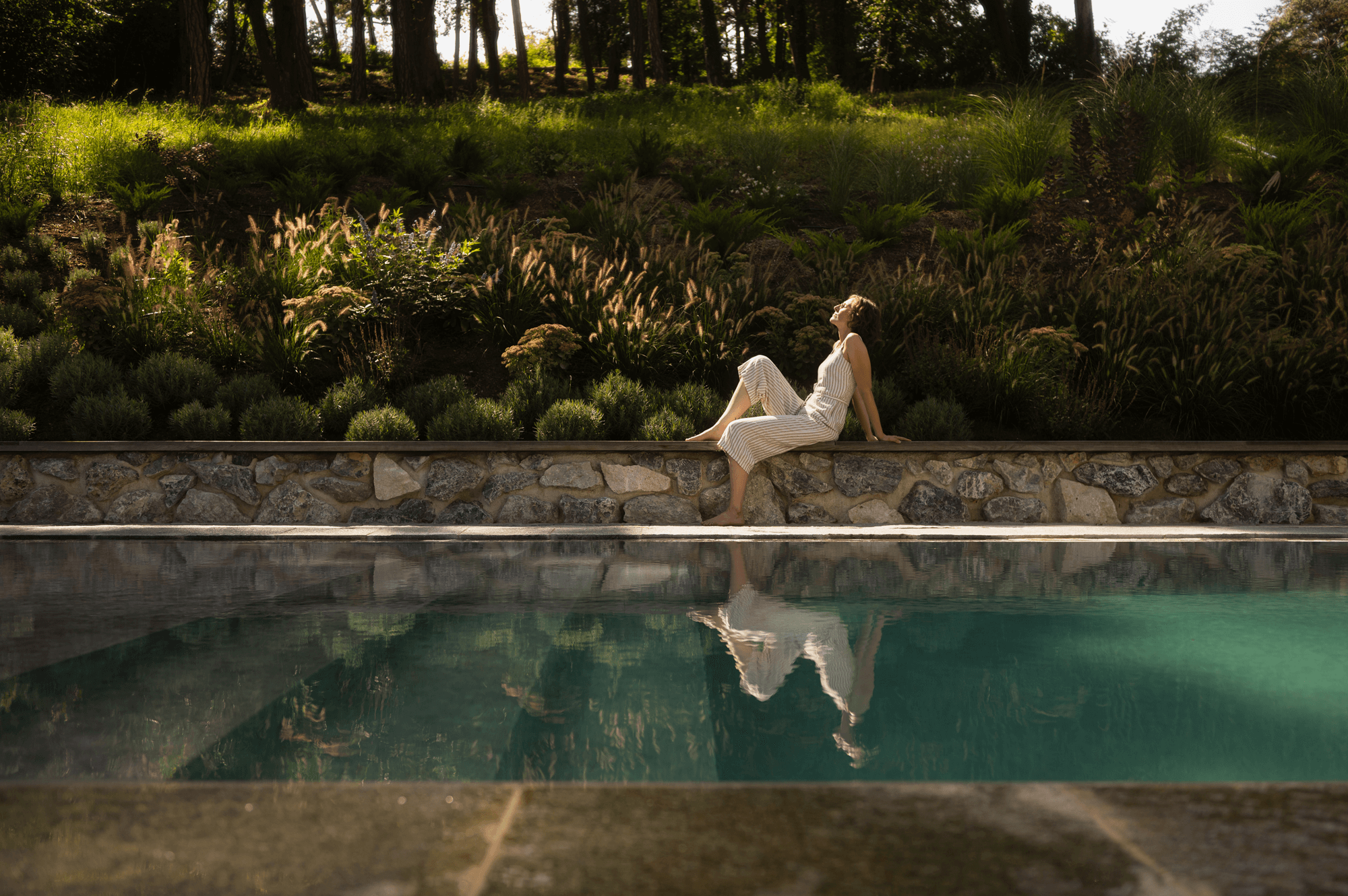 Eine Frau genießt die Sonne neben dem Pool. Im Hintergrund ist ein grün bepflanzter Hang.